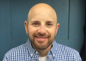 headshot of Jesse. He is a white-appearing bald man with a brown-and-grey short beard, hazel eyes, and gold earrings. He wears a blue and white checked button up shirt.